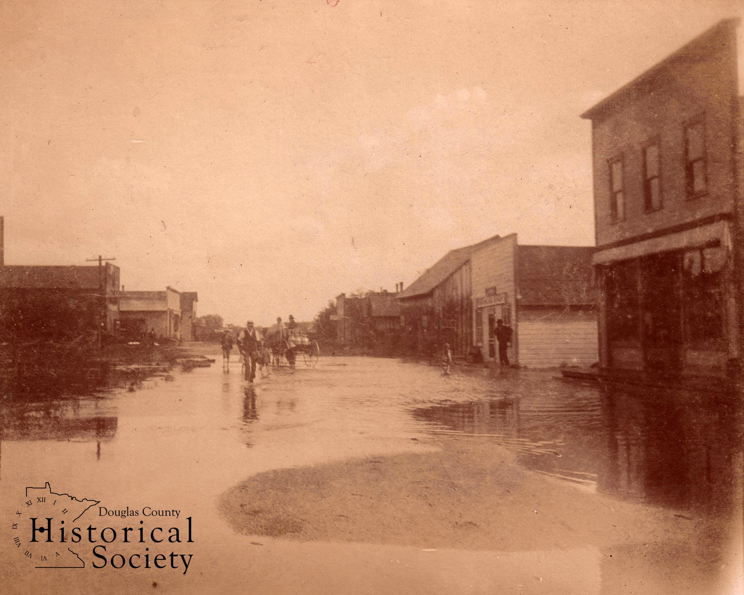 A ca. 1910 photograph of Nelson, showing floodwaters stretching across the entire width of Main Street as people try to navigate in boots, atop wagons, or barefoot while carrying shoes